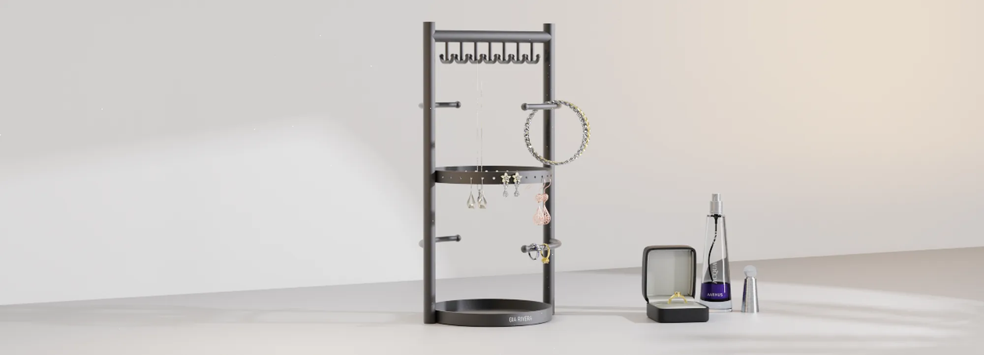 A compact rotating matte black steel Gia Rivera jewellery stand on a round tray base, styled with rings, a bracelet, drop earrings and a necklace against a soft studio backdrop, a ring box and scent bottle beside it, the Gia Rivera wordmark engraved on the tray.