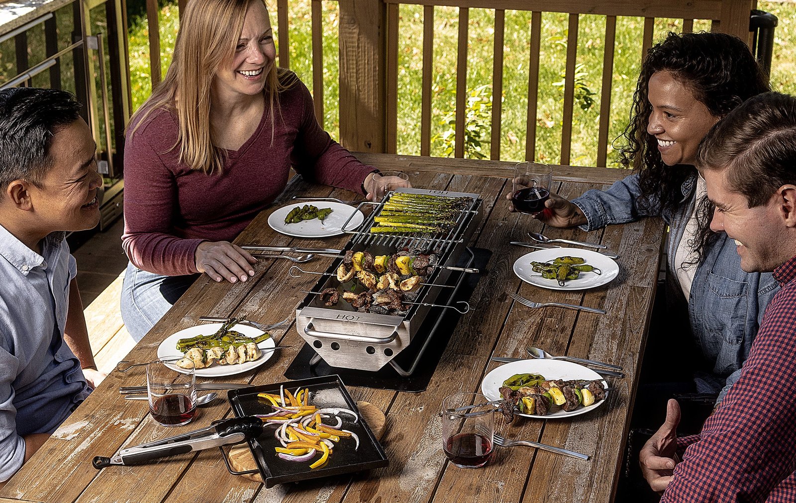 Four friends gathered around an outdoor dining table, the Hickory Summit hibachi grill at the centre with skewers, grilled asparagus and vegetables on its grates and flattop, wine glasses and plates around it.
