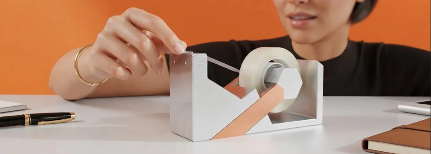 Mono tape dispenser on a desk, a hand pulling a strip of tape from the brushed aluminium body with its anodised accent arm.